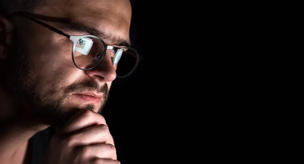 Close-up of a focused poker player with glasses looking at a computer screen in a dark room, analyzing opponents.