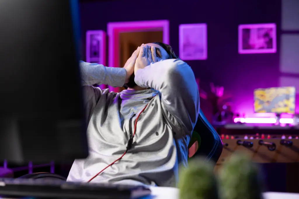 A frustrated online poker player covering his face with his hands in front of a computer monitor, experiencing tilt during a downswing.